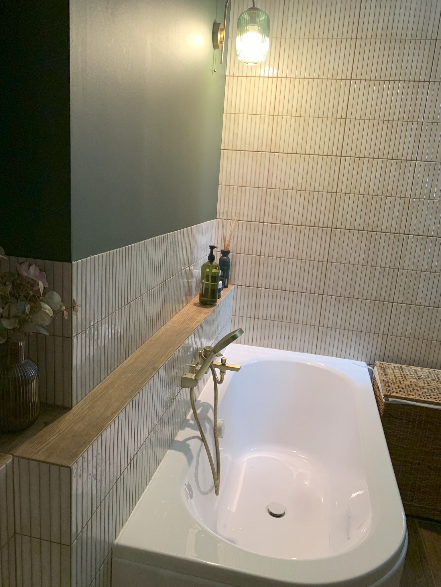 Minimalistyczna łazienka: biała wanna z złotą armaturą, półka z kosmetykami, pionowe płytki, zielona ściana i lampa. Wystrój wnętrza w spokojnych barwach.