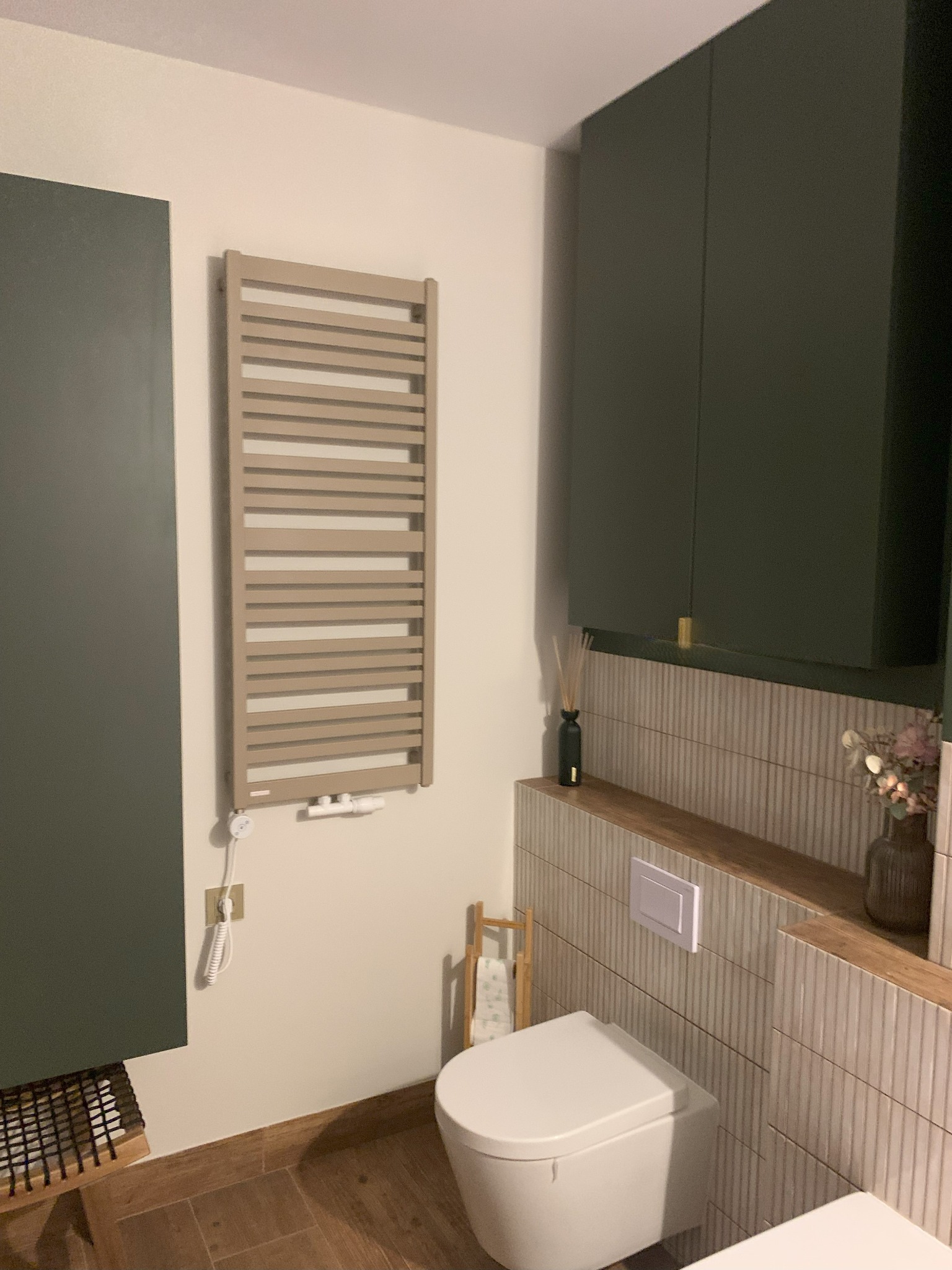 Nowoczesna łazienka: beżowy grzejnik drabinkowy, toaleta z ukrytą spłuczką, zielone szafki wiszące i dekoracyjne płytki w pionowe paski. Minimalistyczny design.