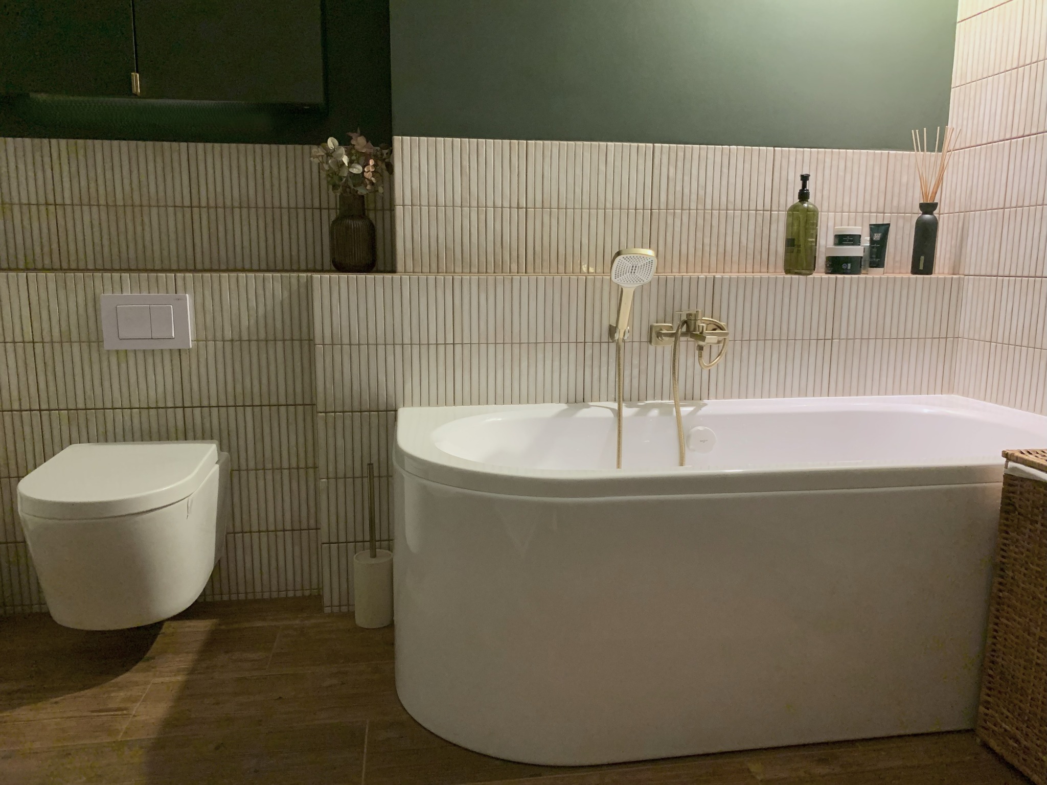 Elegancka łazienka z białą wanną, złotą armaturą i beżowymi płytkami. Na półce dekoracje i kosmetyki. Ciemnozielona ściana nadaje wnętrzu nowoczesny charakter.