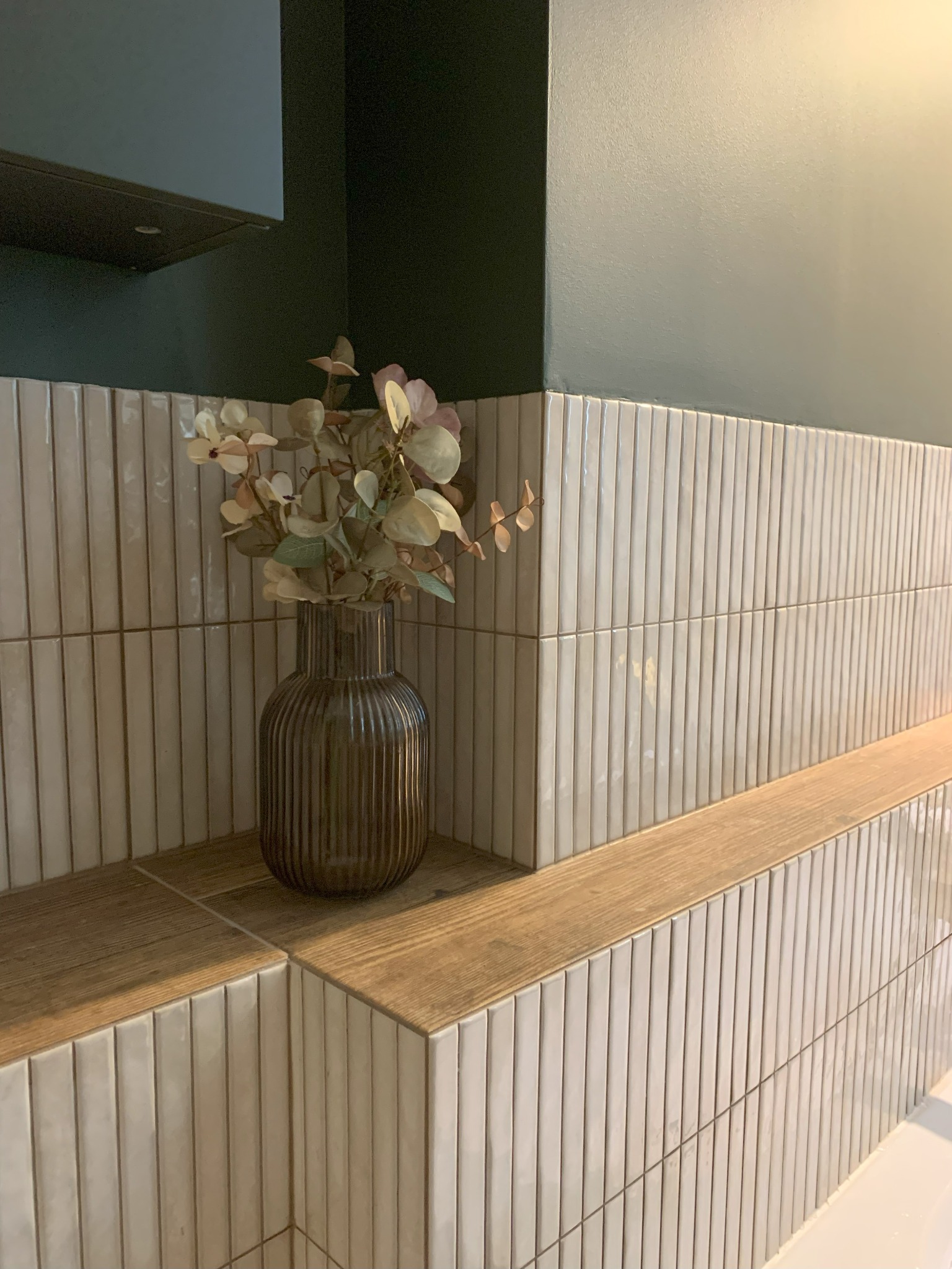 Wazon z suszem na drewnianej półce, na tle ściany z beżowych, wąskich płytek. Górna część ściany pomalowana na ciemnozielony kolor.