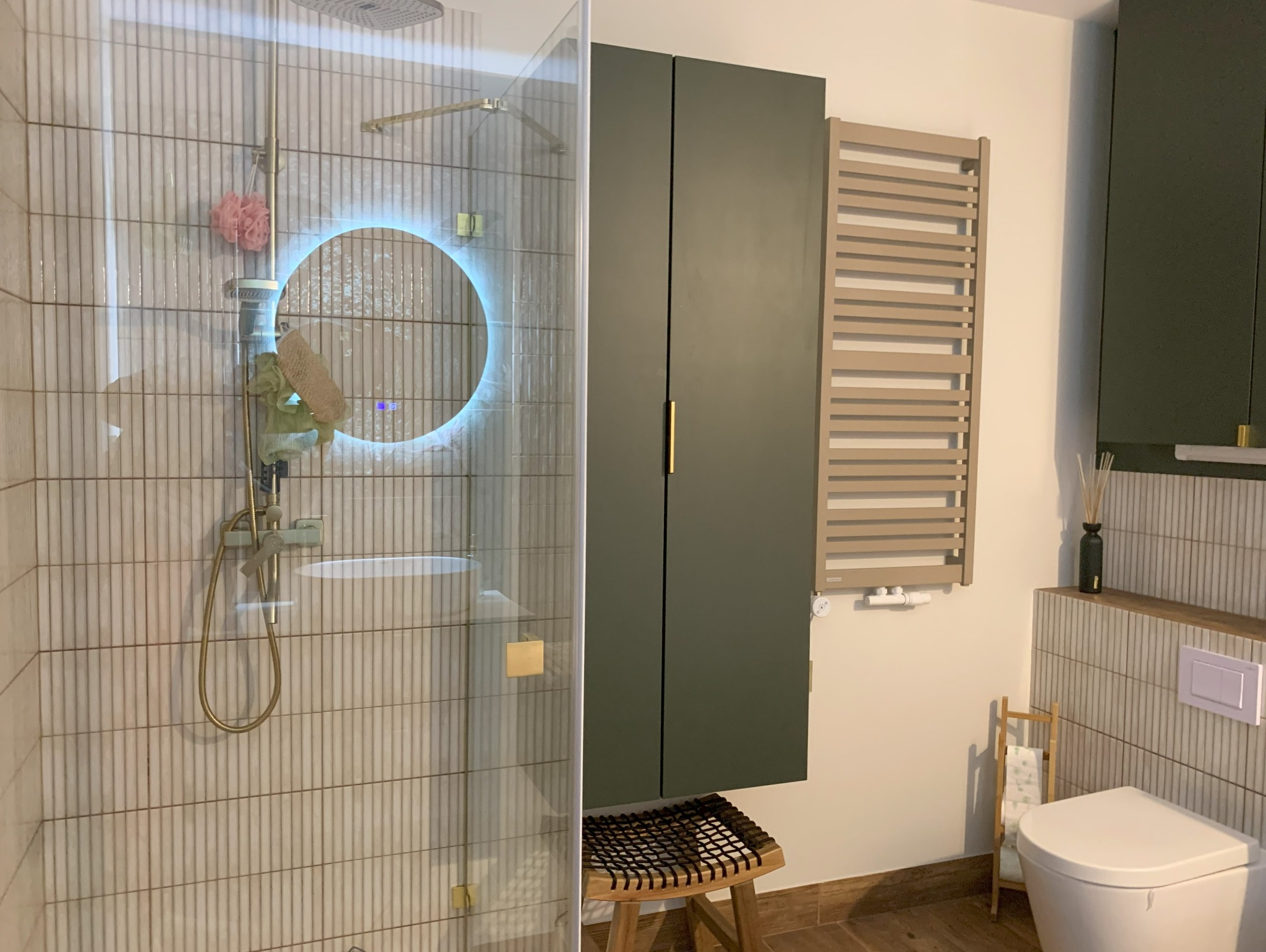 Nowoczesna łazienka z kabiną prysznicową, okrągłym lustrem z podświetleniem LED, grzejnikiem drabinkowym i szafką w kolorze butelkowej zieleni.