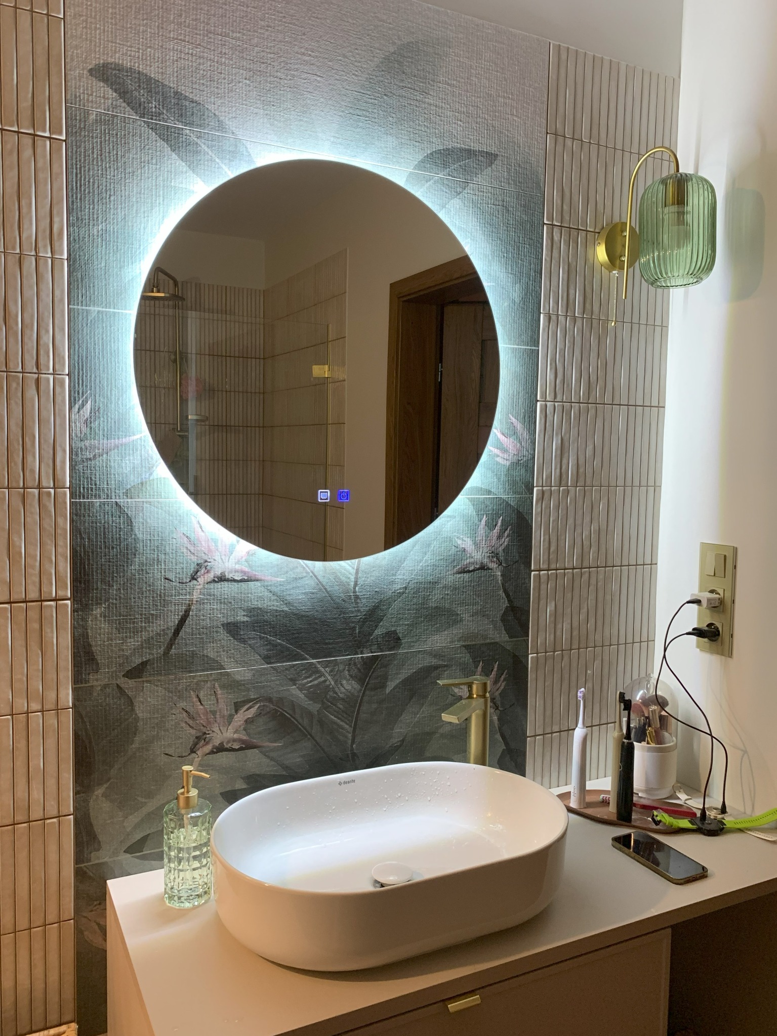 Nowoczesna łazienka z okrągłym lustrem LED, umywalką nablatową i zielonymi akcentami. Widoczne płytki z motywem roślinnym i elegancka lampa ścienna.