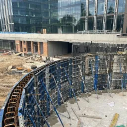Budowa fundamentów o nietypowym, zakrzywionym kształcie, z widocznym szalunkiem i zbrojeniem, na tle nowoczesnego budynku biurowego w trakcie budowy.