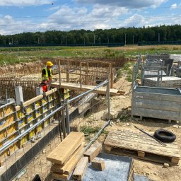 BitTom - Pracownik w kasku i kamizelce odblaskowej pracuje na drewnianym pomoście przy szalunkach fundamentowych na placu budowy domu murowanego, widoczne zbrojenia, palety i narzędzia.