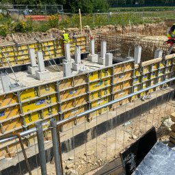 BitTom - Przygotowanie fundamentów pod budynek: szalunki Doka z żółtymi panelami, zalane betonem słupy, widoczny pracownik w kamizelce odblaskowej i kasku, zbrojenie, teren budowy z roślinnością w tle.