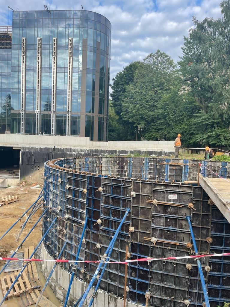 Budowa fundamentów o zakrzywionym kształcie, widoczne stalowe szalunki i podpory, w tle nowoczesny budynek biurowy z przeszkloną fasadą, dwóch pracowników w odblaskowych kamizelkach.