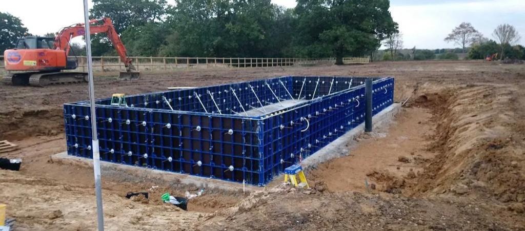 Fundament z szalunków modułowych w kolorze niebieskim, przygotowany do zalania betonem, widoczna koparka w tle.
