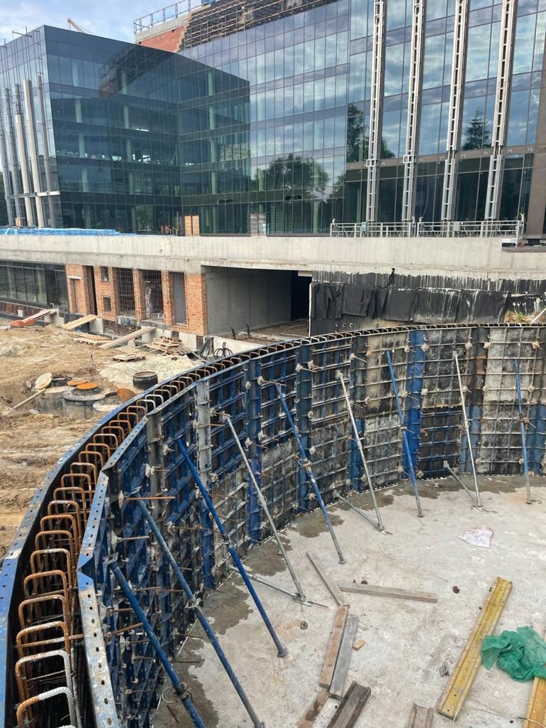 Budowa fundamentów o nietypowym, zakrzywionym kształcie, z widocznym szalunkiem i zbrojeniem, na tle nowoczesnego budynku biurowego w trakcie budowy.