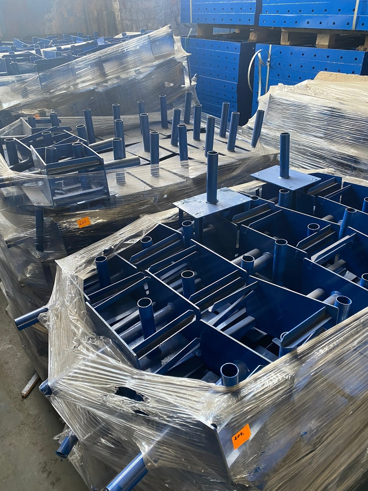 Stosy niebieskich, metalowych elementów szalunkowych do betonu, owiniętych folią stretch z pomarańczowymi naklejkami identyfikacyjnymi, składowane w magazynie.