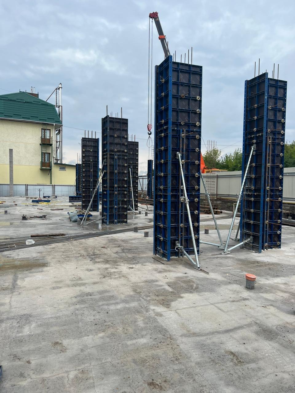 Wznoszenie szkieletu budynku z użyciem dźwigu: widoczne stalowe szalunki kolumnowe, zbrojenie i mokry betonowy fundament w tle budynku mieszkalnego z zielonym dachem.