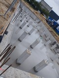 Zalany betonem fundament z wystającymi słupkami i prętami zbrojeniowymi, widoczne szalunki i przygotowany teren budowy w tle.