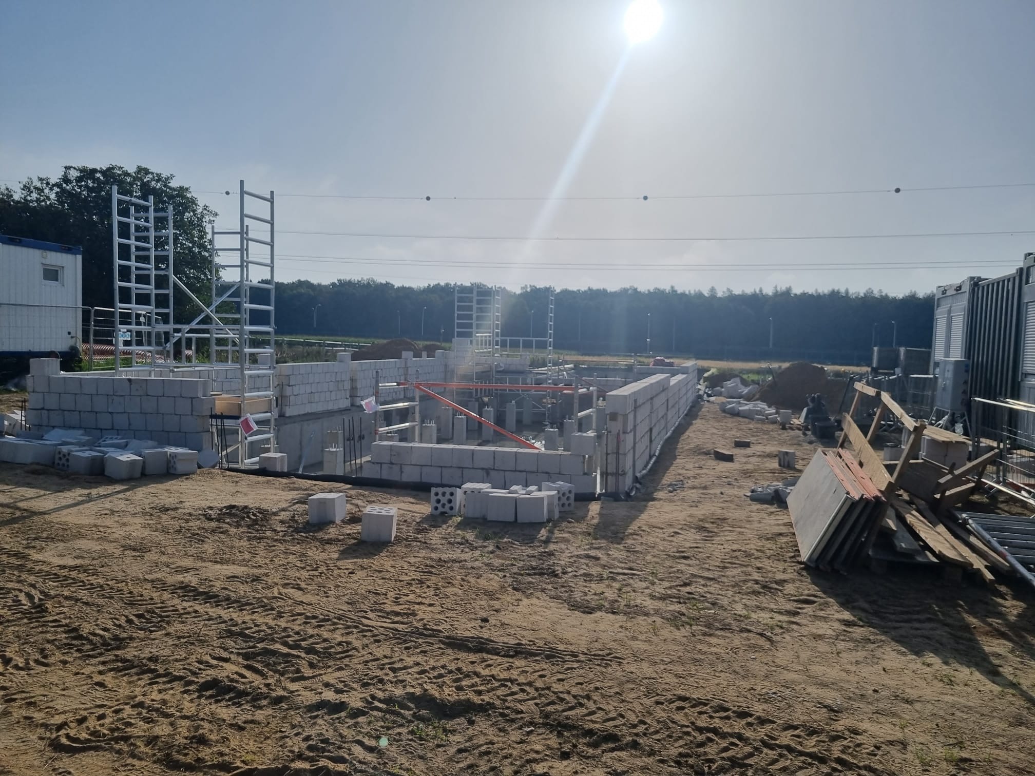 Wznoszenie fundamentów i ścian domu z bloczków betonowych na piaszczystym terenie budowy, widoczne rusztowania i tymczasowe kontenery biurowe w tle, słoneczny dzień.