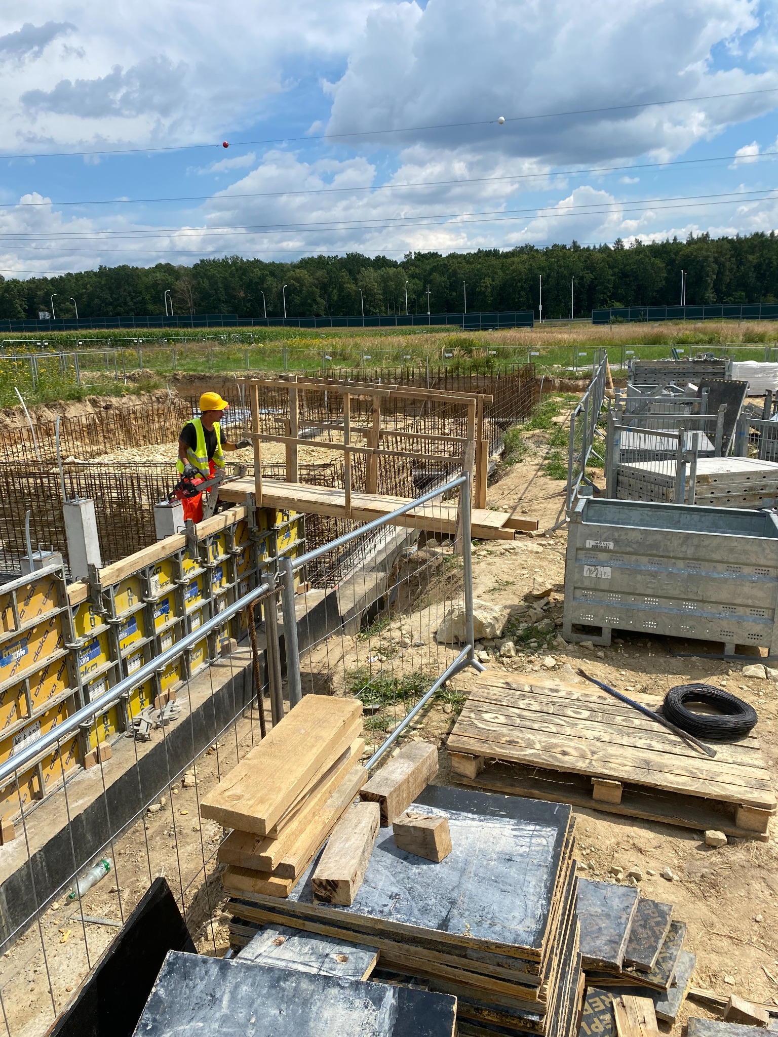 Pracownik w kasku i kamizelce odblaskowej pracuje na drewnianym pomoście przy szalunkach fundamentowych na placu budowy domu murowanego, widoczne zbrojenia, palety i narzędzia.