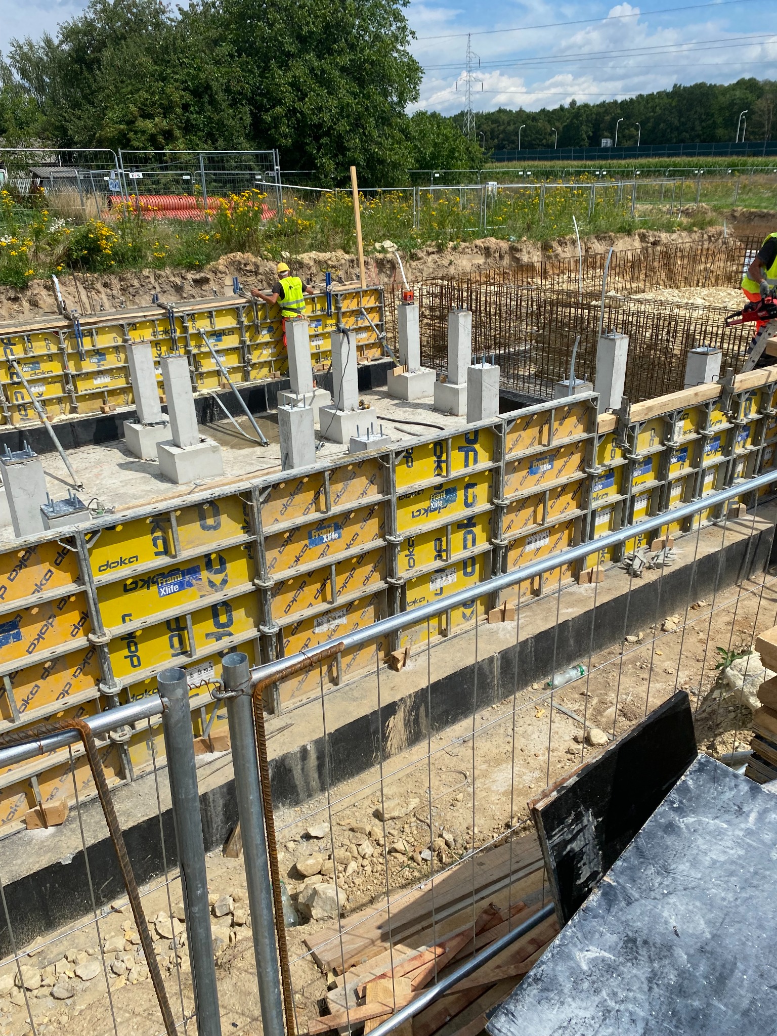 Przygotowanie fundamentów pod budynek: szalunki Doka z żółtymi panelami, zalane betonem słupy, widoczny pracownik w kamizelce odblaskowej i kasku, zbrojenie, teren budowy z roślinnością w tle.