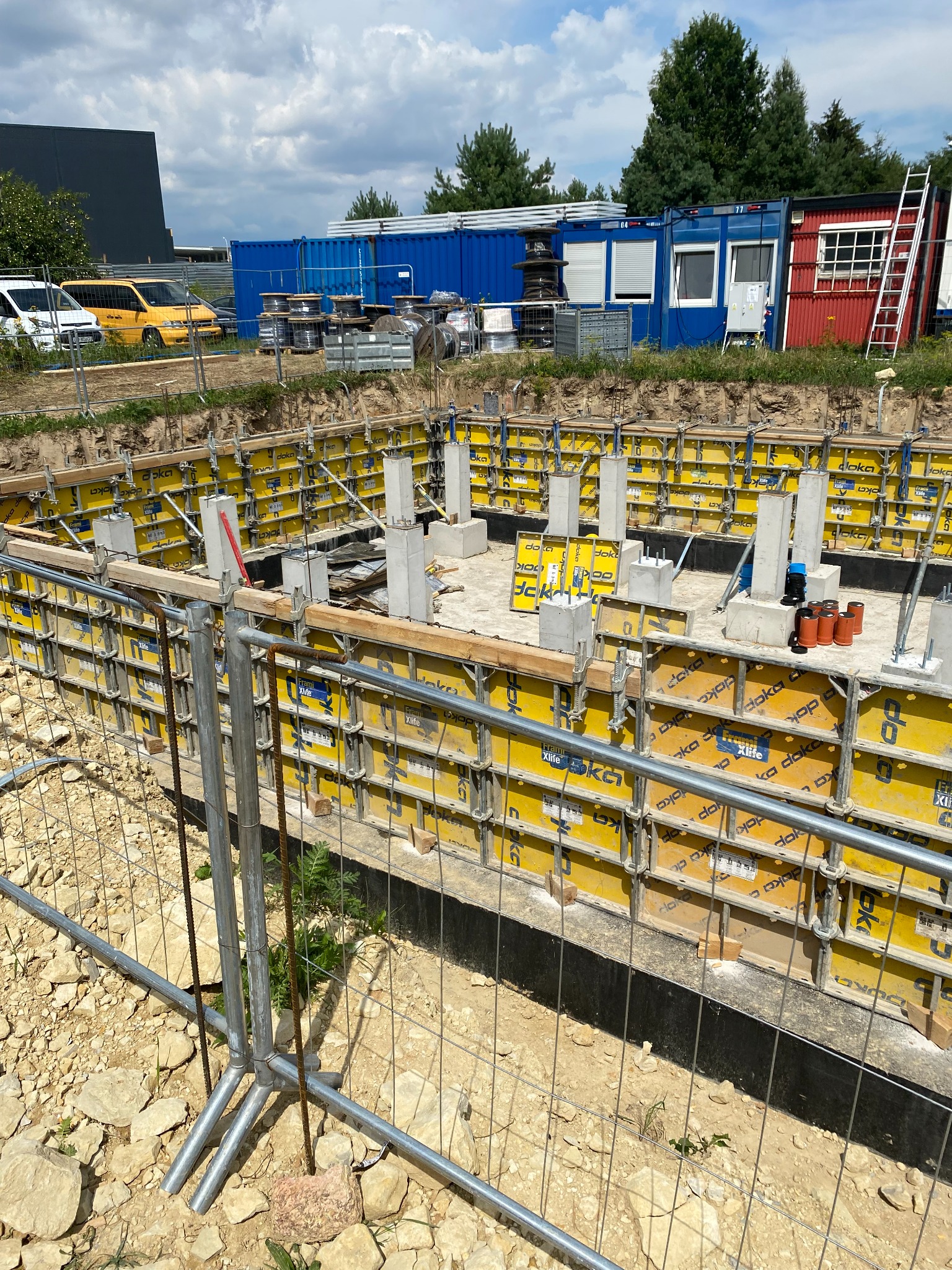 Przygotowanie fundamentów pod budynek: żółte szalunki z betonu, widoczne zbrojenia, tymczasowe podpory i zabezpieczenia, w tle kontenery budowlane i pojazdy.