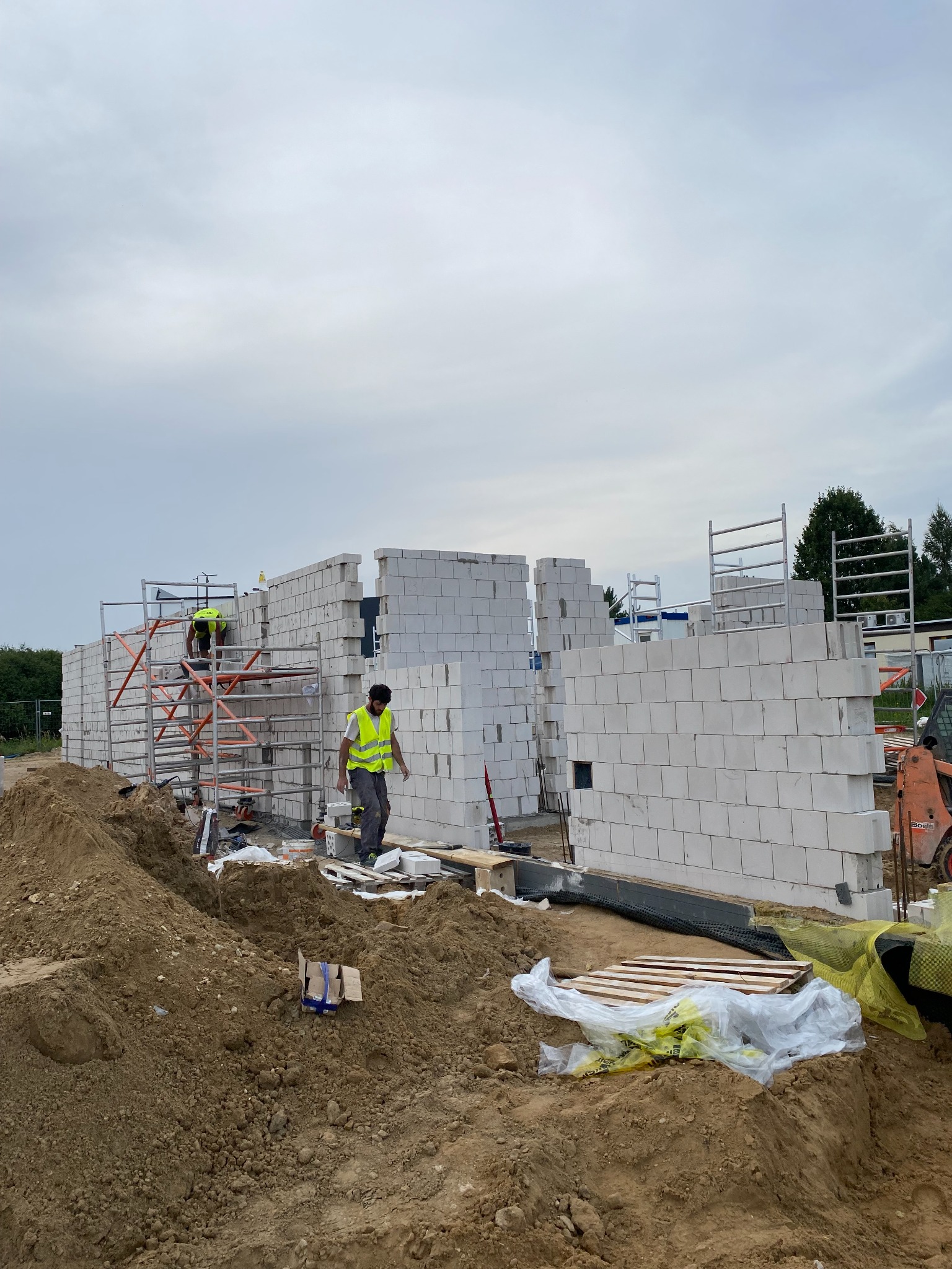 Budowa domu z białych bloczków: pracownicy w kamizelkach odblaskowych na rusztowaniu i jeden na ziemi, widoczny miniładowarka Bobcat, ziemia i materiały budowlane na placu budowy.