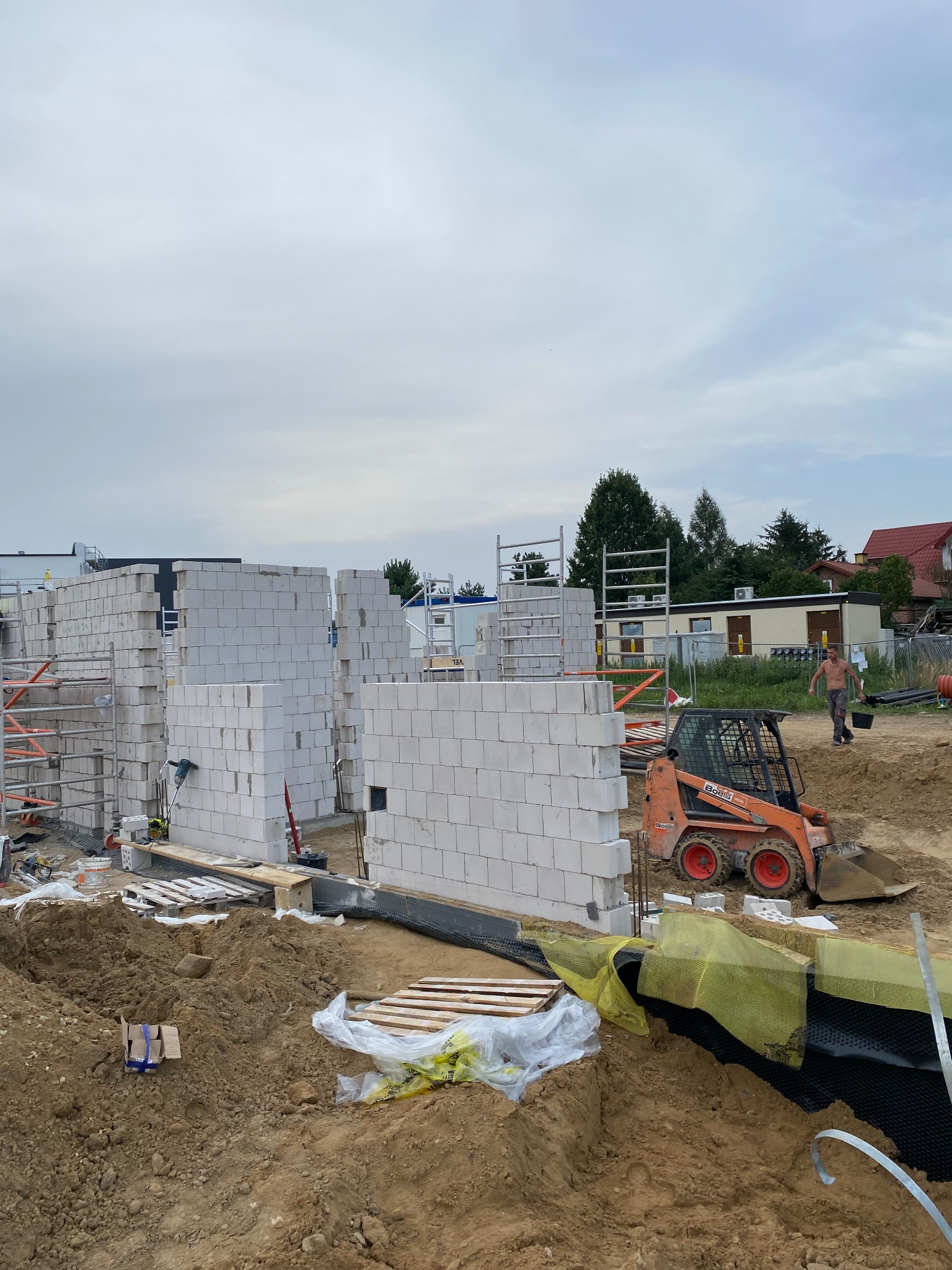 Wznoszenie ścian z bloczków betonu komórkowego Ytong na budowie domu jednorodzinnego, widoczny pomarańczowy miniładowarka Bobcat i pracownik w tle.