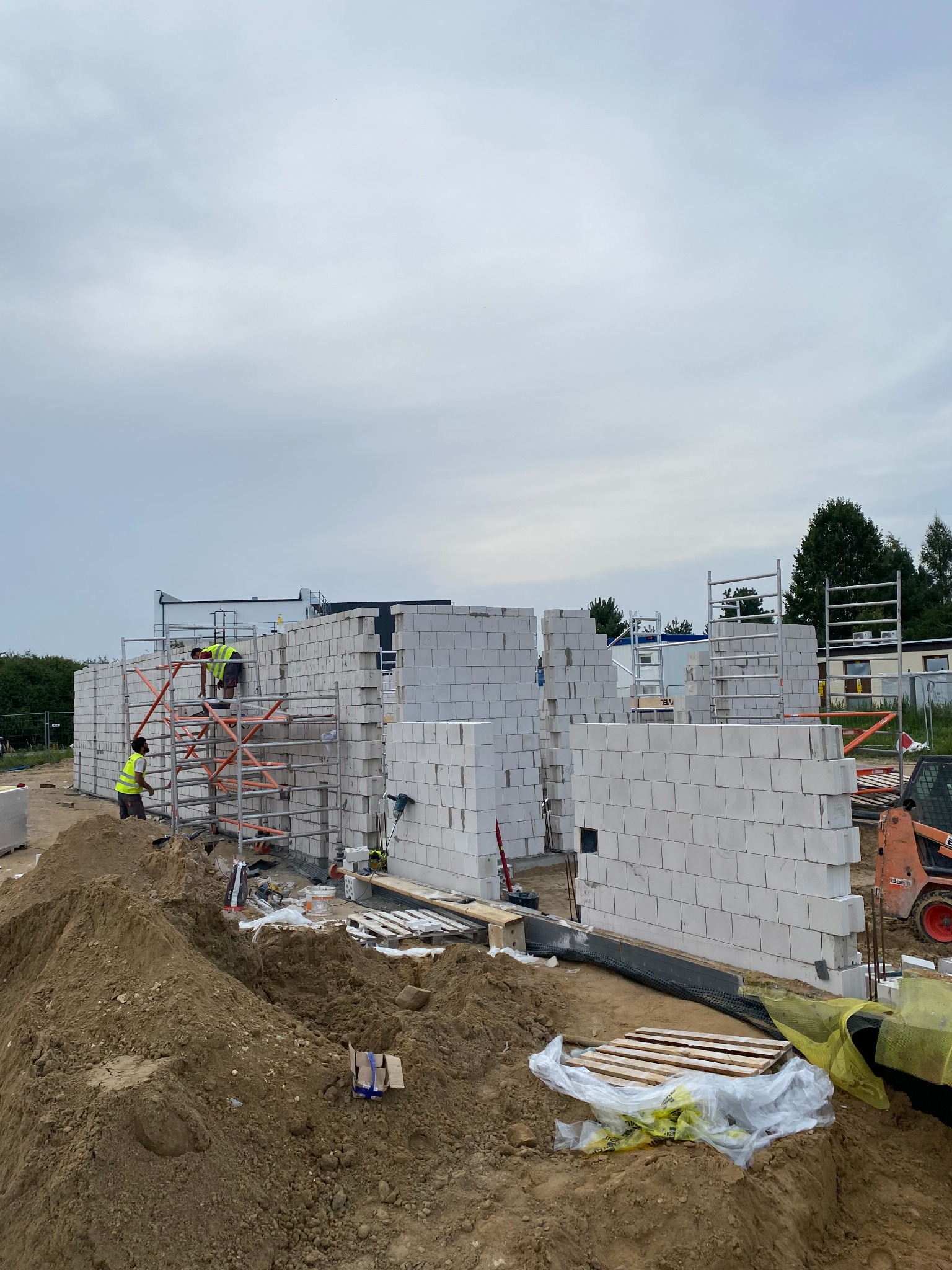 Budowa domu z białych bloczków: dwóch pracowników w żółtych kamizelkach na rusztowaniu, widoczny stos bloczków, koparka i sterta ziemi.