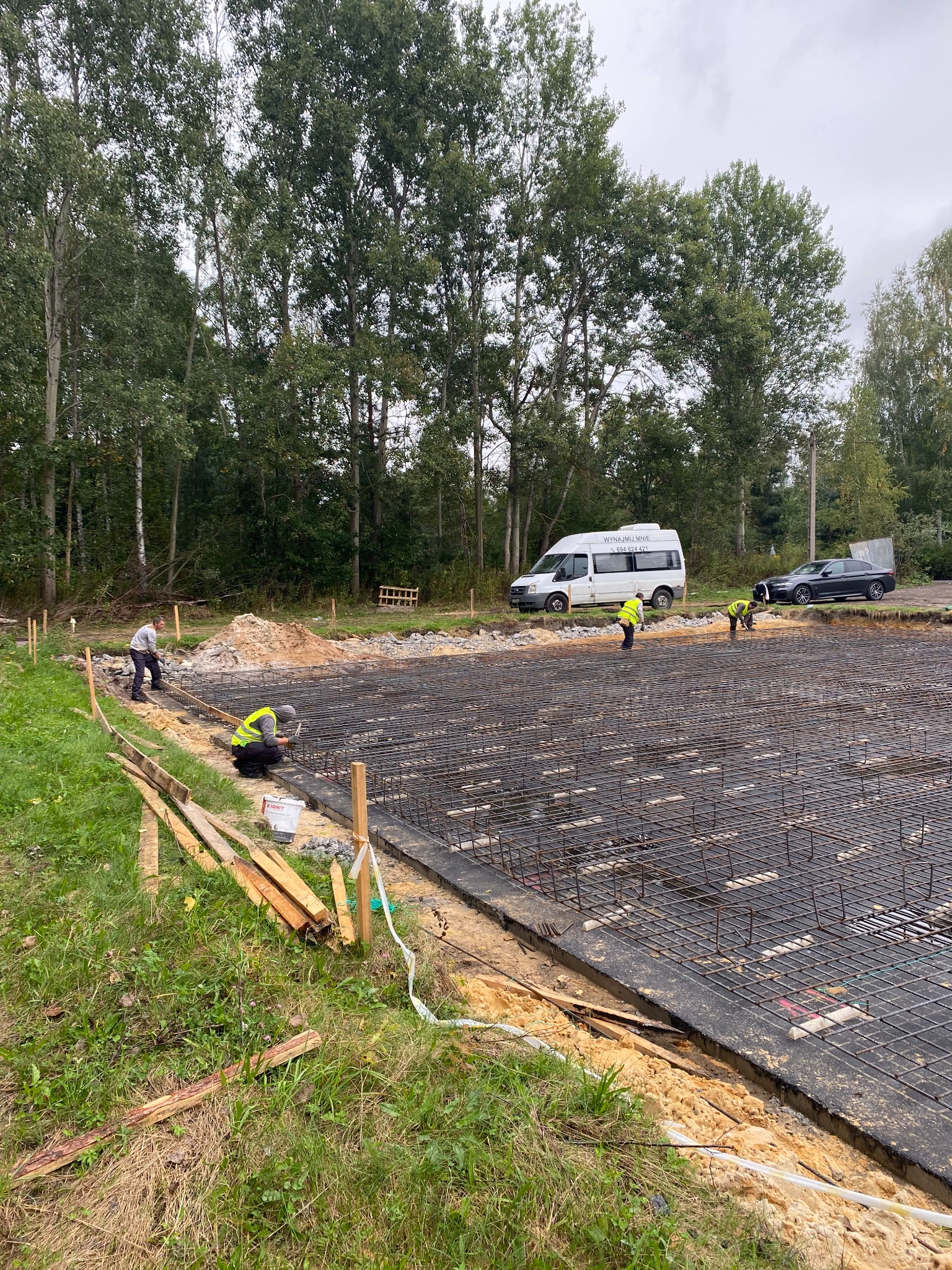 Przygotowanie zbrojenia pod fundament płyty budynku jednorodzinnego, widoczni pracownicy w kamizelkach odblaskowych, z boku leżące deski szalunkowe, w tle samochód dostawczy z numerem telefonu.