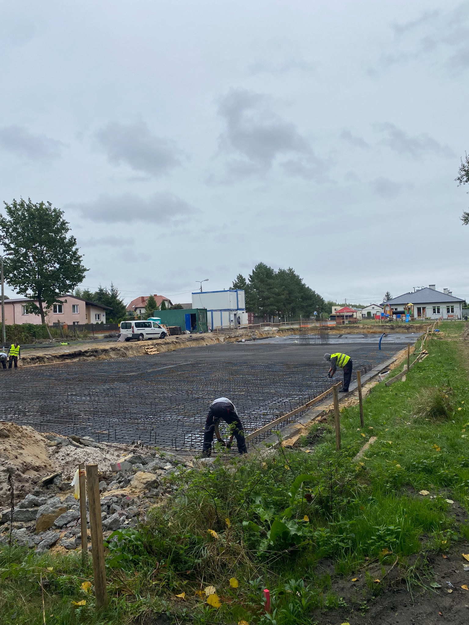 Zbrojenie fundamentów pod budynek jednorodzinny, widoczne pręty zbrojeniowe tworzące siatkę, pracownicy w kamizelkach odblaskowych przy pracy, w tle zabudowa jednorodzinna i pochmurne niebo.