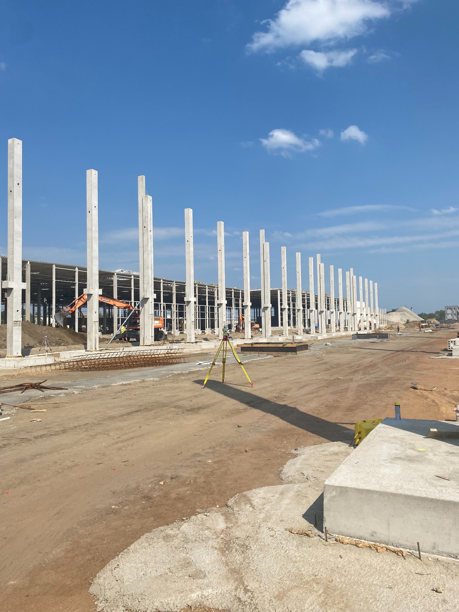 Wznoszenie konstrukcji hali: widok na szkielet budynku z prefabrykatów betonowych, z pracującą koparką w tle i niwelatorem na pierwszym planie, w słoneczny dzień.