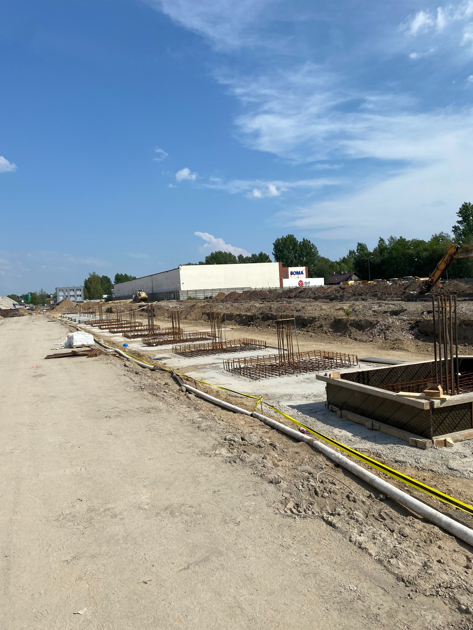 Fundamenty pod budynek komercyjny w trakcie wylewania, widoczne zbrojenia i szalunki, w tle budynek magazynowy z logo firmy.