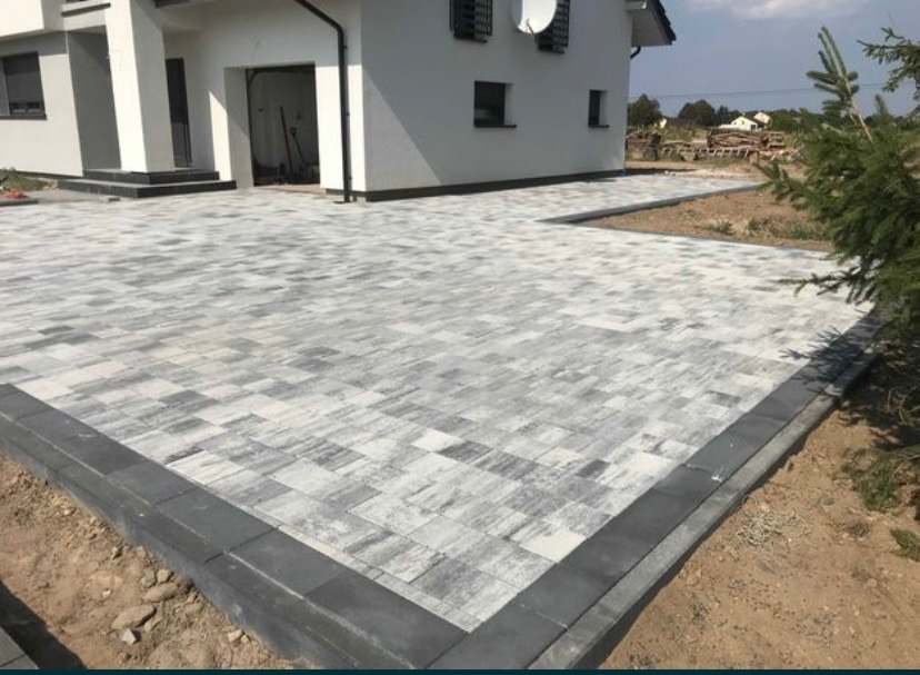 Ułożona kostka brukowa w odcieniach szarości przed nowoczesnym domem z białymi ścianami, widoczne ciemne obrzeża i fragment trawnika.