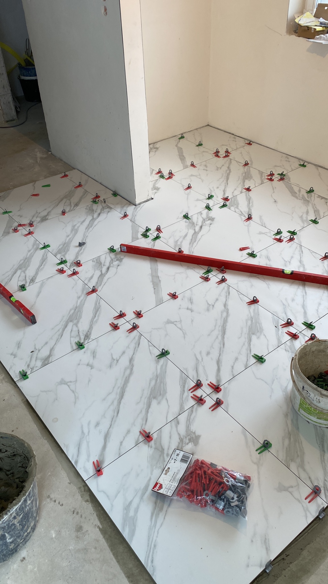 Białe płytki imitujące marmur, wyrównywane za pomocą czerwonych i zielonych klipsów poziomujących, widoczna poziomica i wiadro z zaprawą.
