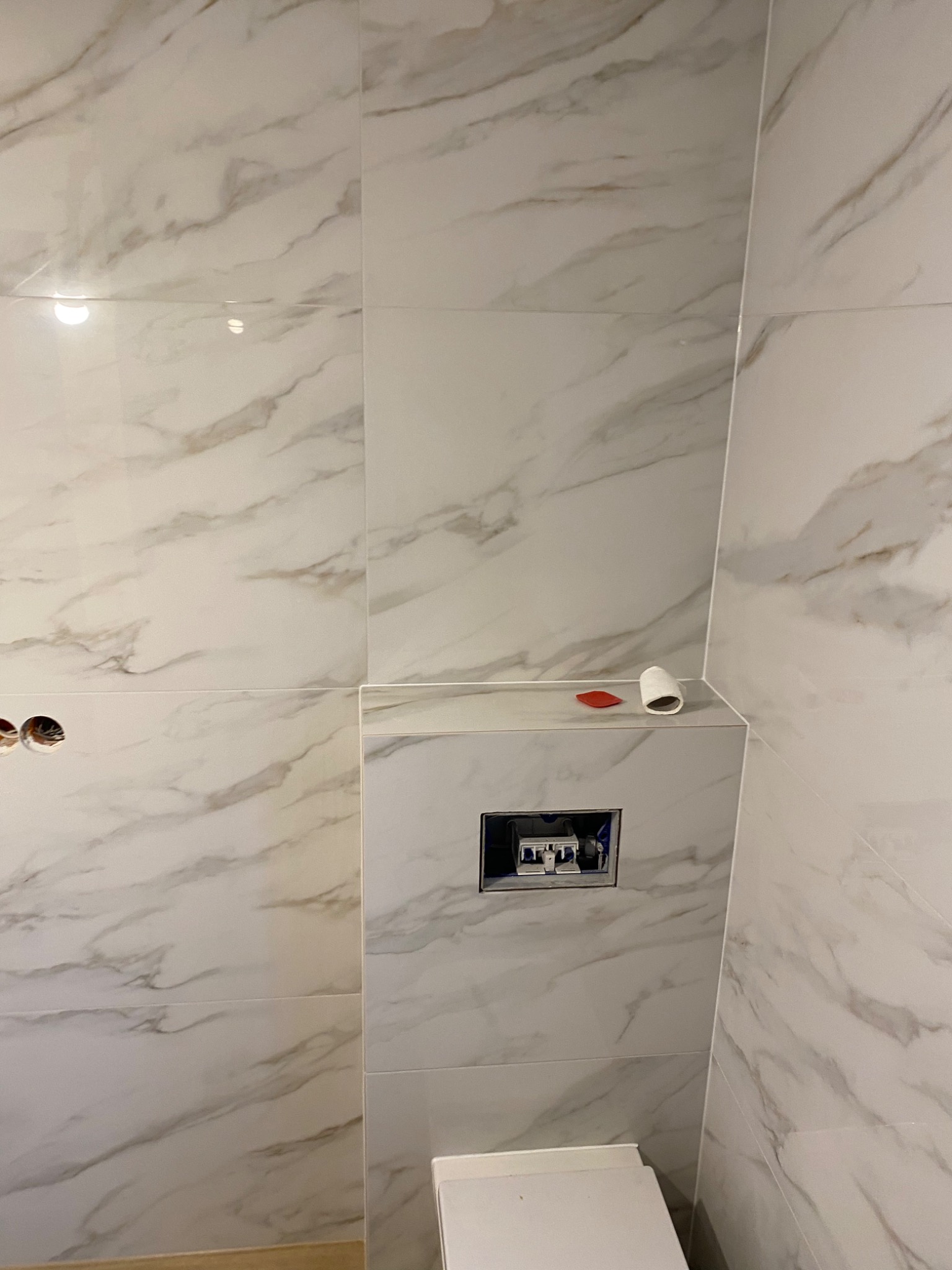 Ściana łazienki wyłożona marmurkowymi płytkami, widoczne otwory na gniazdka elektryczne, stelaż podtynkowy WC, czerwona szpatułka i rolka papieru toaletowego.