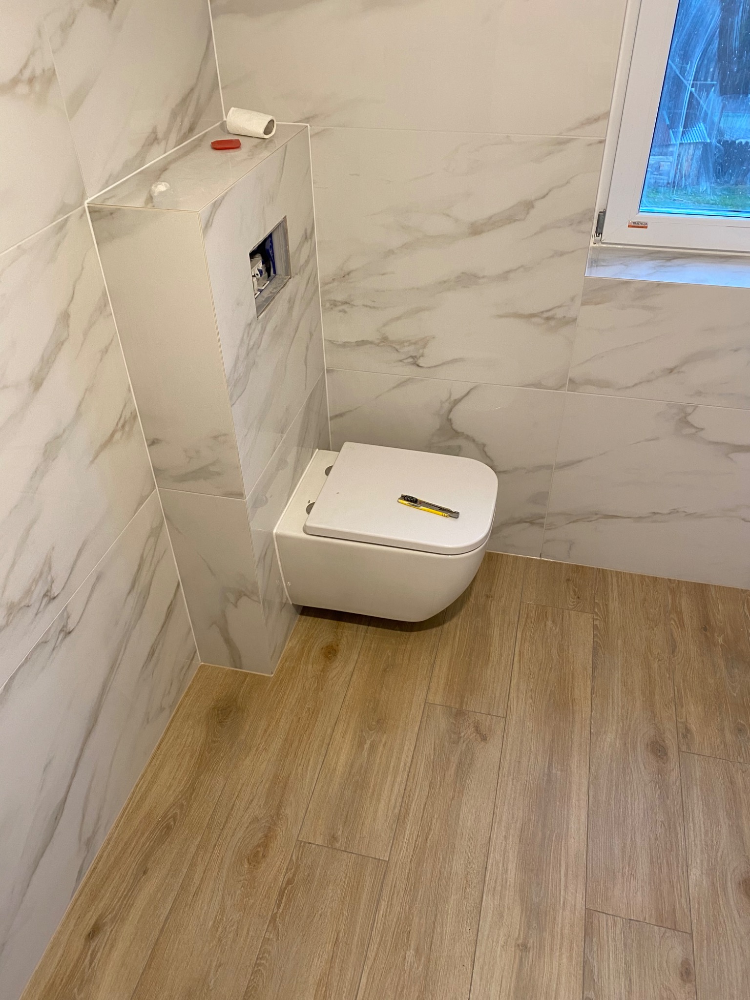 Wisząca toaleta z deską, na której leży nóż do tapet, w łazience wykończonej marmurowymi płytkami i drewnopodobną podłogą, z widocznym oknem w tle.