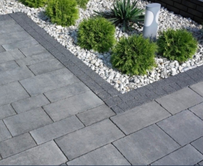 Szara kostka brukowa ułożona wzorem prostokątnym, obrzeża z ciemniejszej kostki, rabata z białym kamieniem i krzewami w tle.