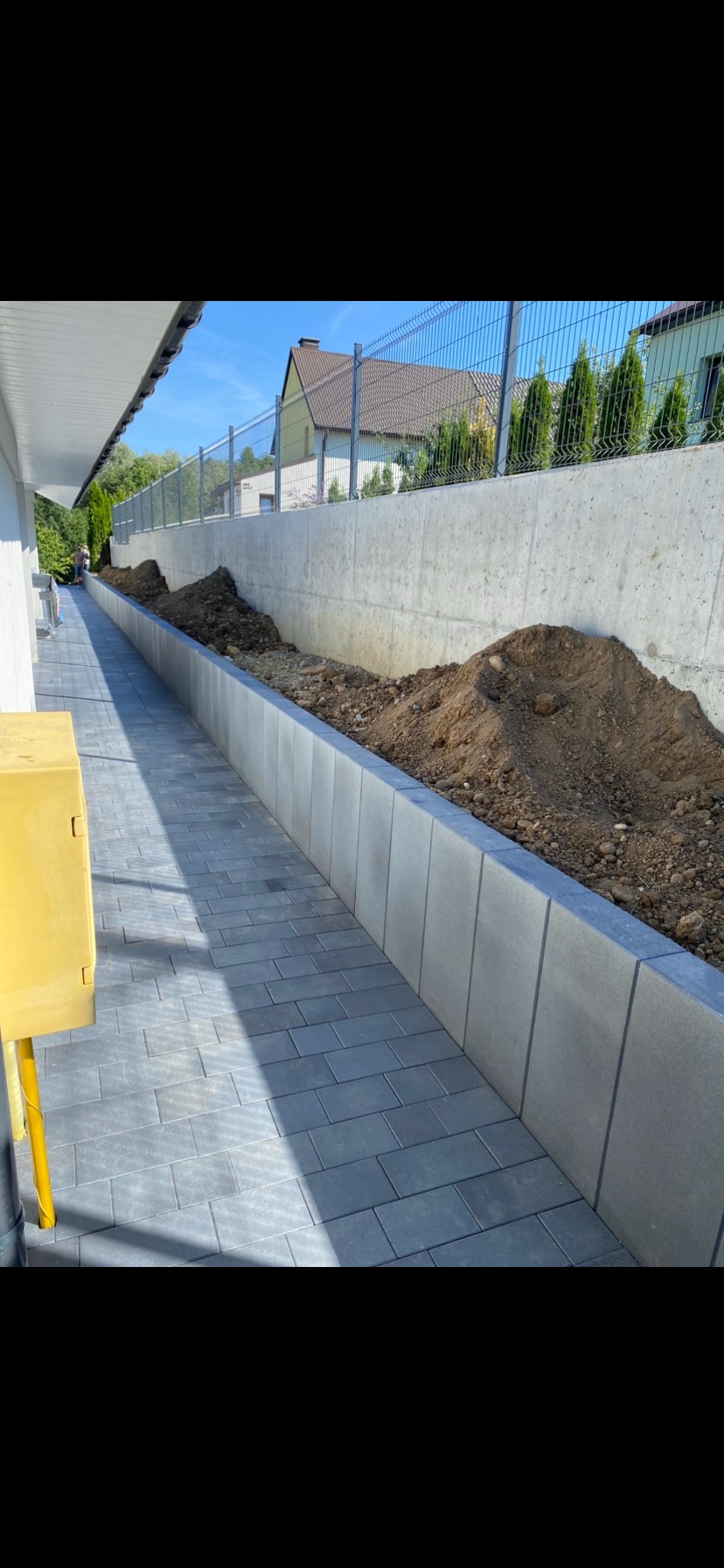 Ułożona szara kostka brukowa tworząca ścieżkę wzdłuż betonowego murku oporowego z nasypaną ziemią, widok na dom z metalową siatką ogrodzeniową w tle, słoneczny dzień.