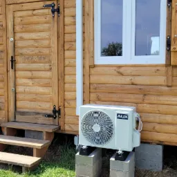 Zewnętrzna jednostka klimatyzacji AUX zamontowana na betonowych bloczkach przy drewnianym domku, z widoczną instalacją odprowadzającą skropliny i fragmentem okna z odbiciem nieba.