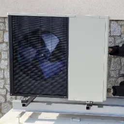 Zewnętrzna jednostka pompy ciepła zamontowana na metalowej konstrukcji przy ścianie z kamienia, widoczne izolowane rury.