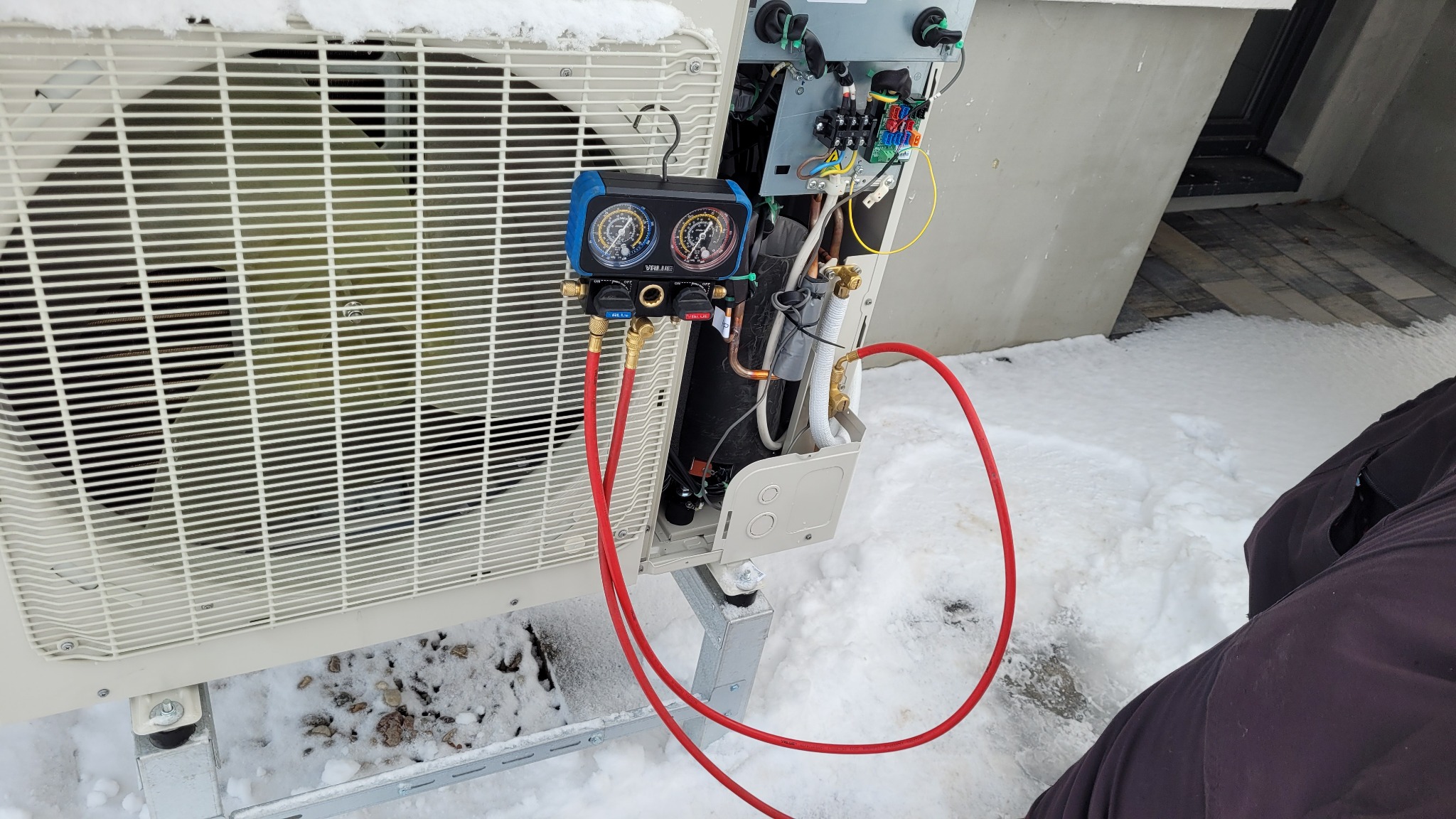 Zewnętrzna jednostka urządzenia z podłączonymi manometrami serwisowymi, widoczna instalacja elektryczna i rurki miedziane, śnieg na obudowie i wokół urządzenia.