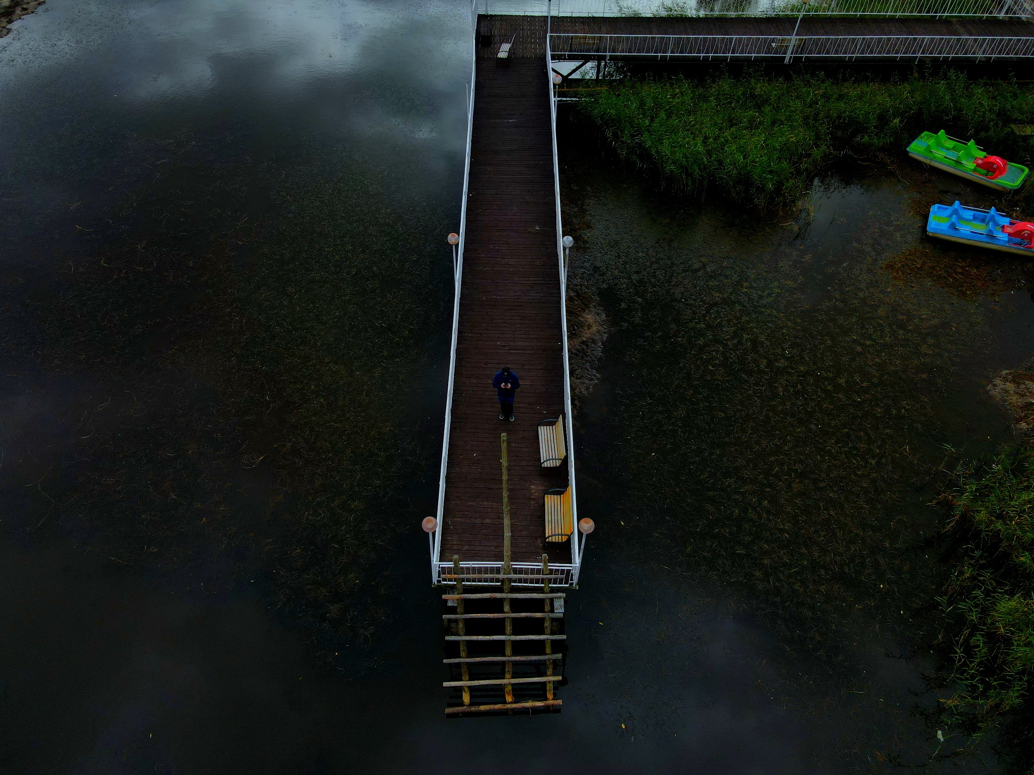 Widok z góry na drewniany pomost z osobą na końcu, otoczony ciemną wodą i zielenią, z kolorowymi rowerkami wodnymi w tle.