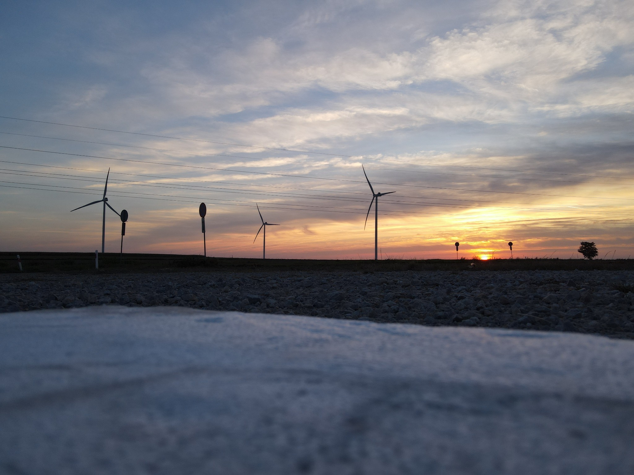 Zachód słońca nad farmą wiatrową widziany z perspektywy niskiego kąta, z betonową krawędzią w pierwszym planie i sylwetkami turbin wiatrowych na tle pomarańczowego nieba.