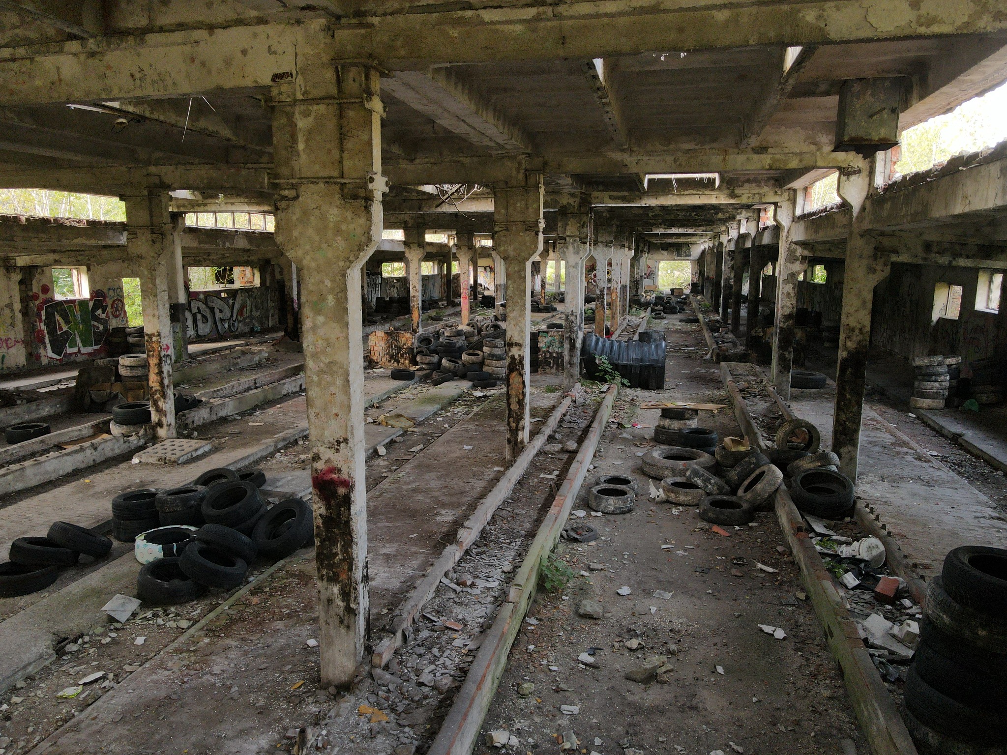 Wnętrze opuszczonej hali produkcyjnej z betonowymi kolumnami i stertami opon, z graffiti na ścianach w tle.