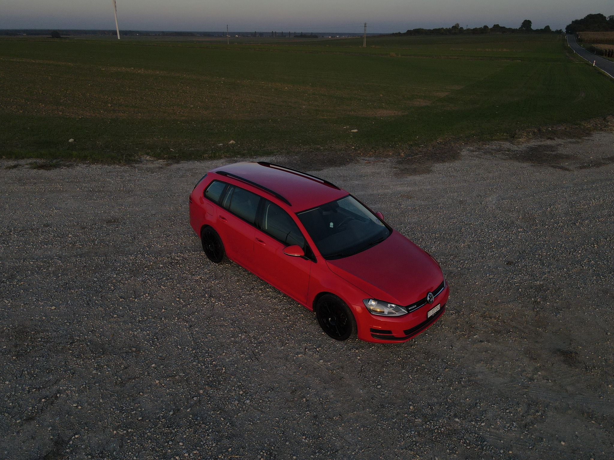 Czerwony samochód kombi z czarnymi felgami, sfotografowany z góry na żwirowym podłożu, z polami i drogą w tle.