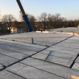 Montaż prefabrykowanych płyt stropowych na budowie domu, widoczny dźwig i pracownik w oddali.