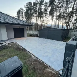 Ułożona kostka brukowa na podjeździe przed domem z szarym garażem blaszanym, widoczne elementy ogrodzenia i przygotowane płyty do dalszych prac.