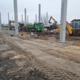 Budowa hali: widok na plac budowy z maszynami (koparki, wywrotka) i wzniesionymi betonowymi słupami konstrukcyjnymi, na ziemi widoczne ślady opon.