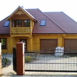 Żółty dom z bali z czerwonym dachem, balkonem i dwoma bramami garażowymi, ogrodzony kutym płotem z ceglanymi słupkami.