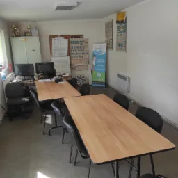 Sala szkoleniowa z dwoma drewnianymi stołami, krzesłami, komputerami i plakatami ze znakami drogowymi na ścianie. W tle szafa z trofeami i baner reklamowy.