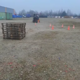 Pomarańczowy wózek widłowy z operatorem, ustawiony na placu manewrowym z pachołkami i paletami, w tle budynek magazynowy i drzewa, pochmurne niebo.