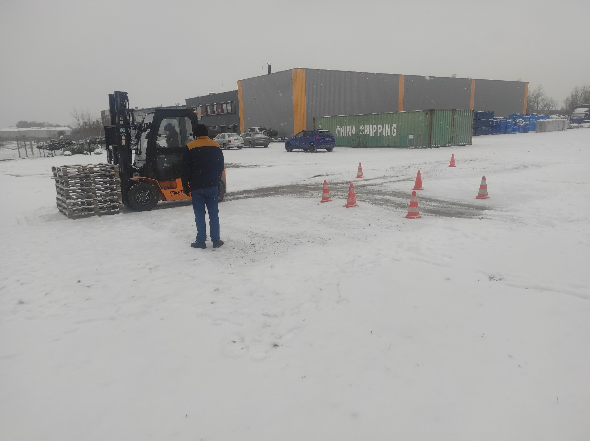 Operator w niebieskich spodniach i kurtce z żółtymi elementami stoi obok wózka widłowego z paletami na śniegu, w tle budynek przemysłowy i pomarańczowe pachołki.