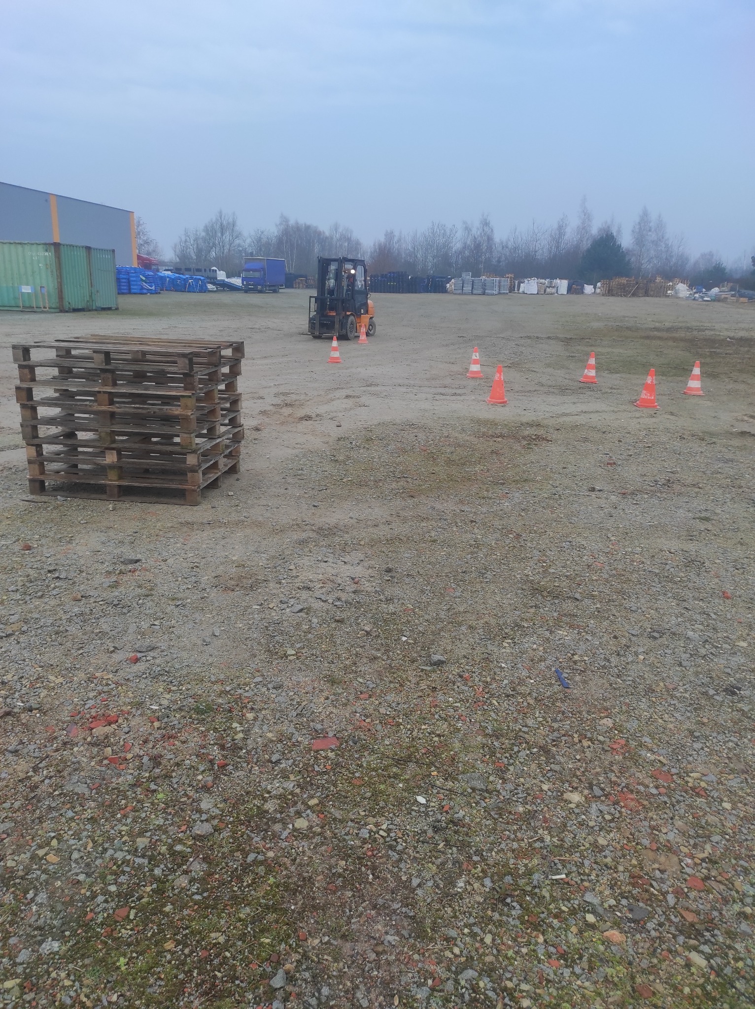 Pomarańczowy wózek widłowy z operatorem, ustawiony na placu manewrowym z pachołkami i paletami, w tle budynek magazynowy i drzewa, pochmurne niebo.