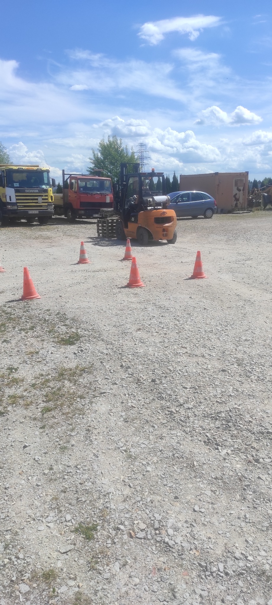 Pomarańczowa ładowarka teleskopowa na placu manewrowym z paletą i pachołkami, w tle zaparkowane samochody i ciężarówki.