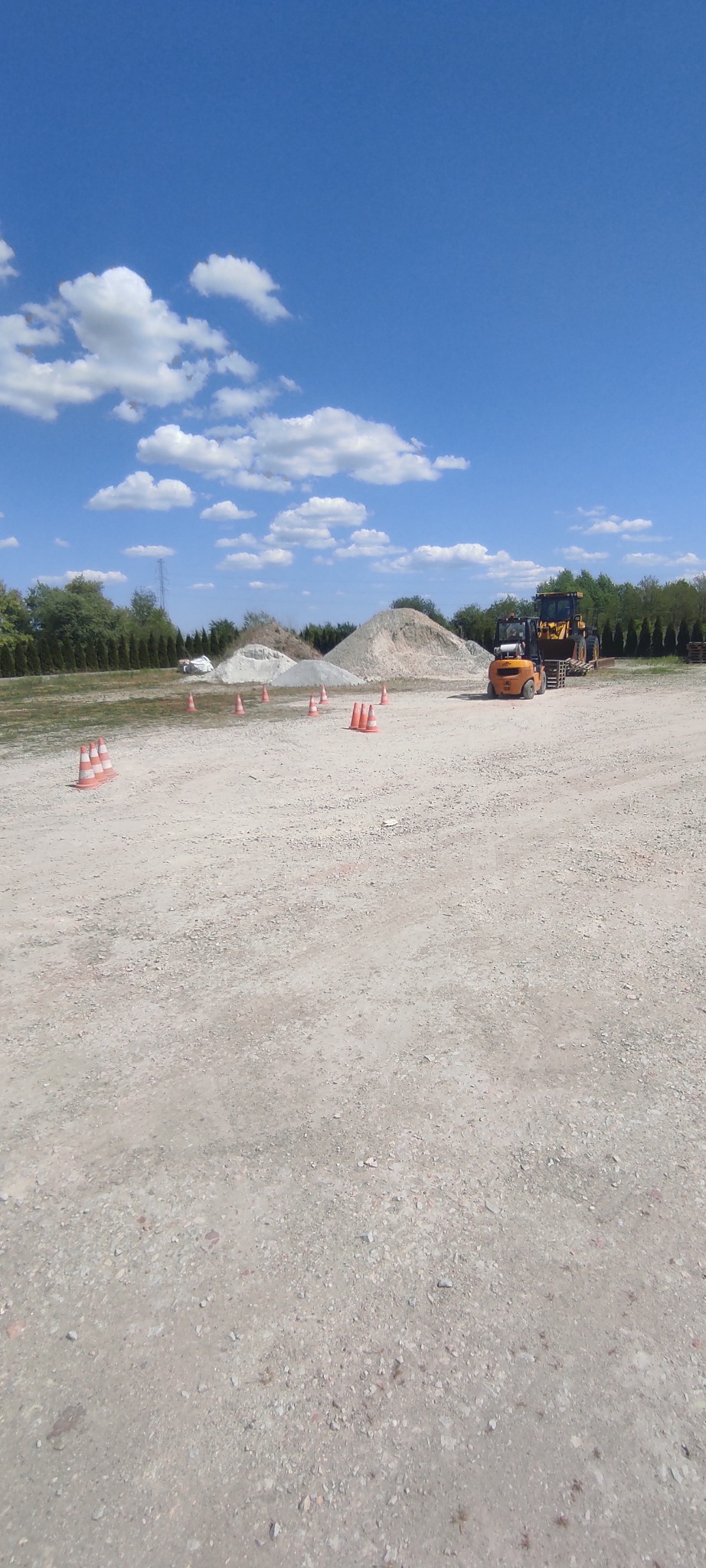 Żółta ładowarka teleskopowa na lawecie transportowej, ustawiona na żwirowym placu z pryzmami piasku i wygrodzona pomarańczowymi pachołkami, błękitne niebo z chmurami w tle.