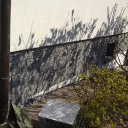 Szara elewacja budynku z imitacji cegły, widoczna rynna, cień gałęzi na ścianie i krzew z młodymi pędami